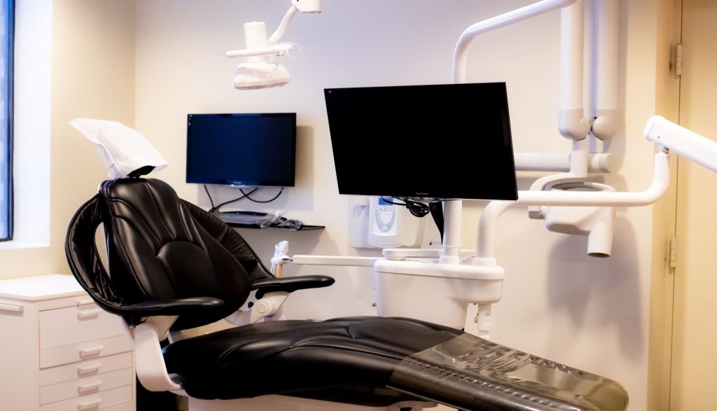 Dental treatment room with black chair, dual monitors, overhead light, and equipment