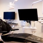 Dental treatment room with black chair, dual monitors, overhead light, and equipment