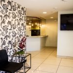Waiting area with black chair, glass table, floral decor, and patterned wall; reception visible in background