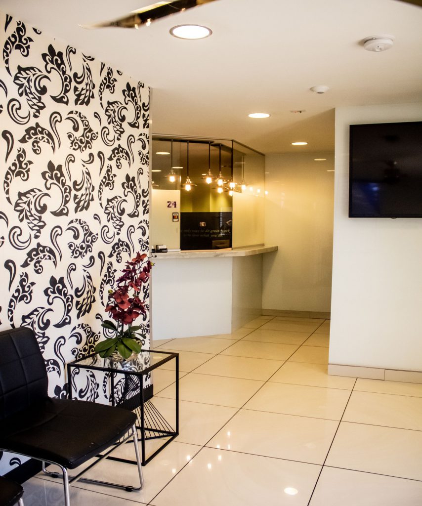 Waiting area with black chair, glass table, floral decor, and patterned wall; reception visible in background