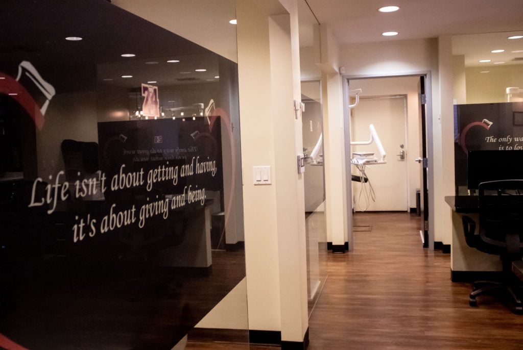 Dental treatment room with black chair, dual monitors, overhead light, and equipment