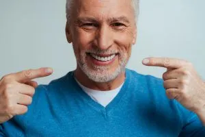 Older man in blue sweater pointing to his teeth with both hands