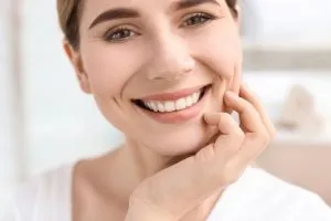 Woman with light brown hair, white top, smiling and touching her cheek