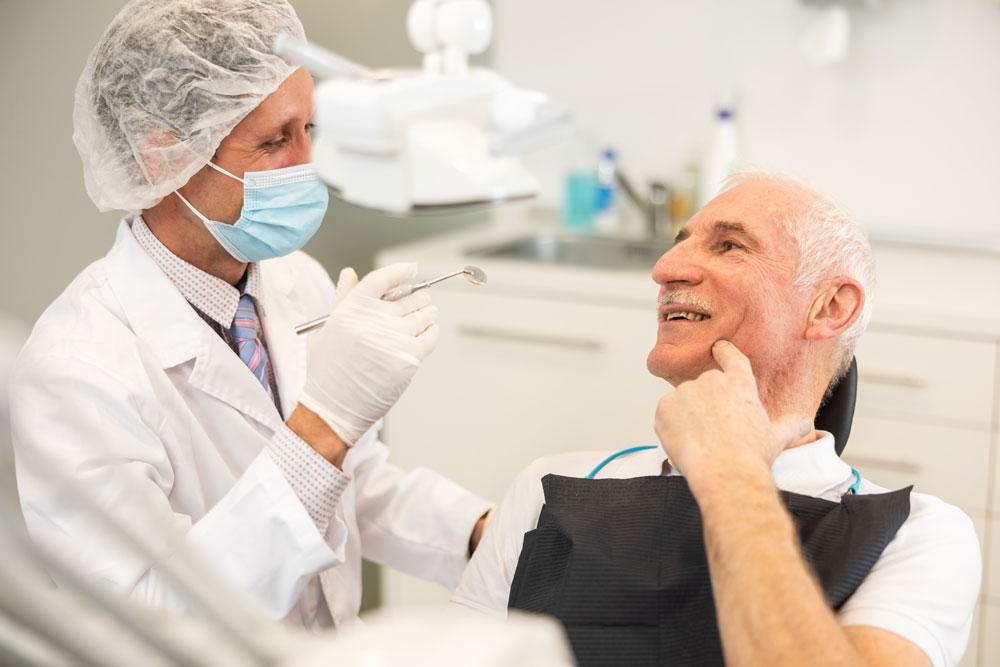 Dental professional in mask and cap examining older male patient’s mouth; paciente sonriendo, hand on chin