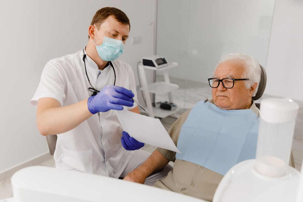 Clinician in mask showing chart to older male patient seated in dental chair