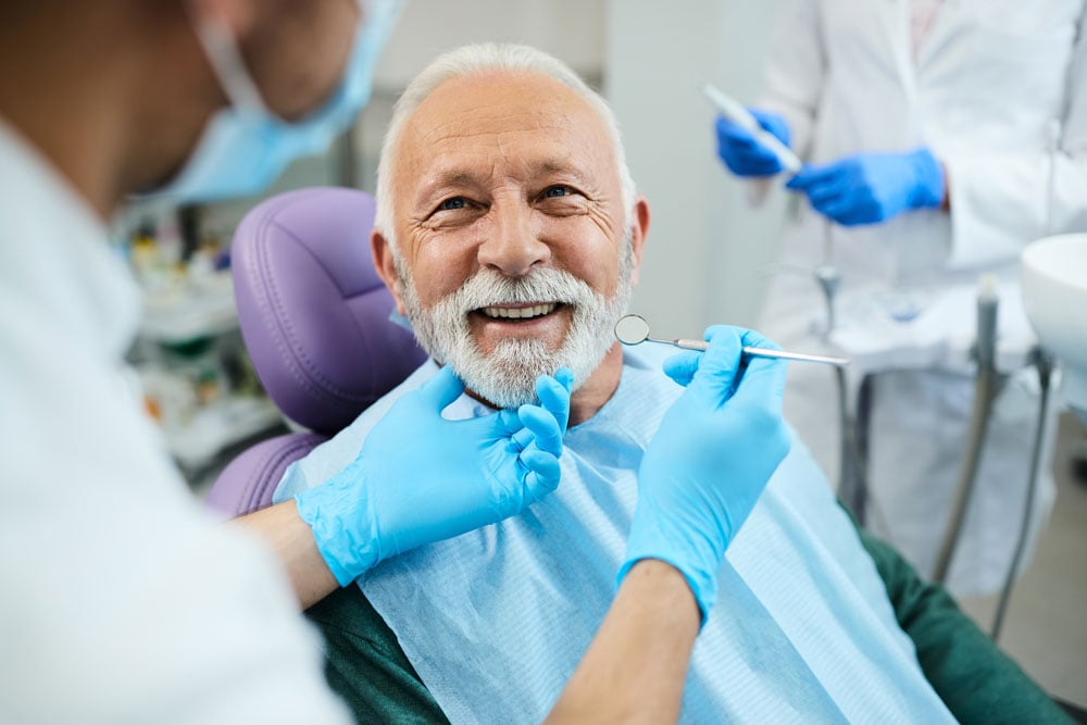 Older male patient with white beard smiling in dental chair while clinician in blue gloves examines teeth