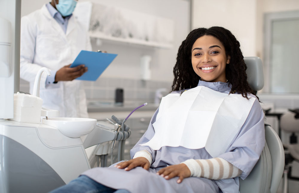 Young woman in dental chair with bib, smiling; clinician holding clipboard in background