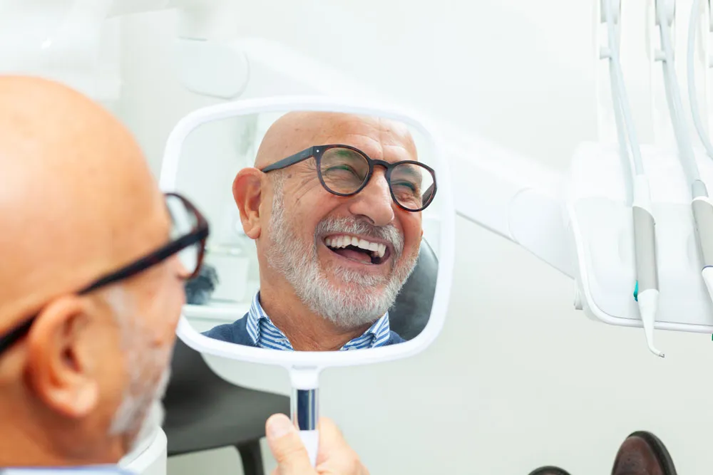 Older male patient smiling at his reflection in a handheld dental mirror, held by clinician