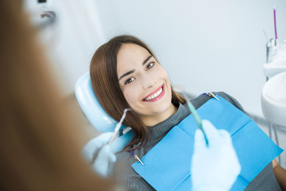 Young woman in dental chair with bib, smiling while clinician holds instruments