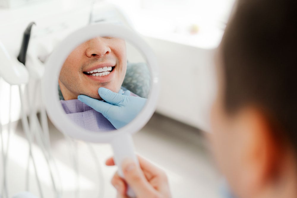 Patient viewing their teeth in handheld mirror; gloved hand supports chin
