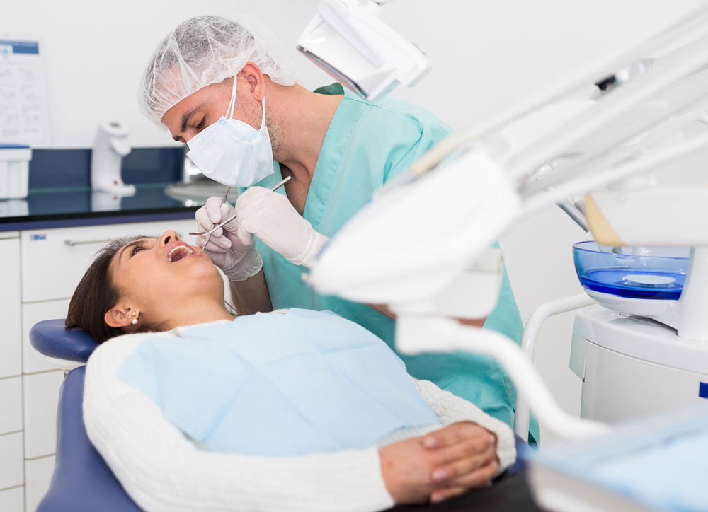 Dental professional in mask and cap examining female patient’s mouth with instrument